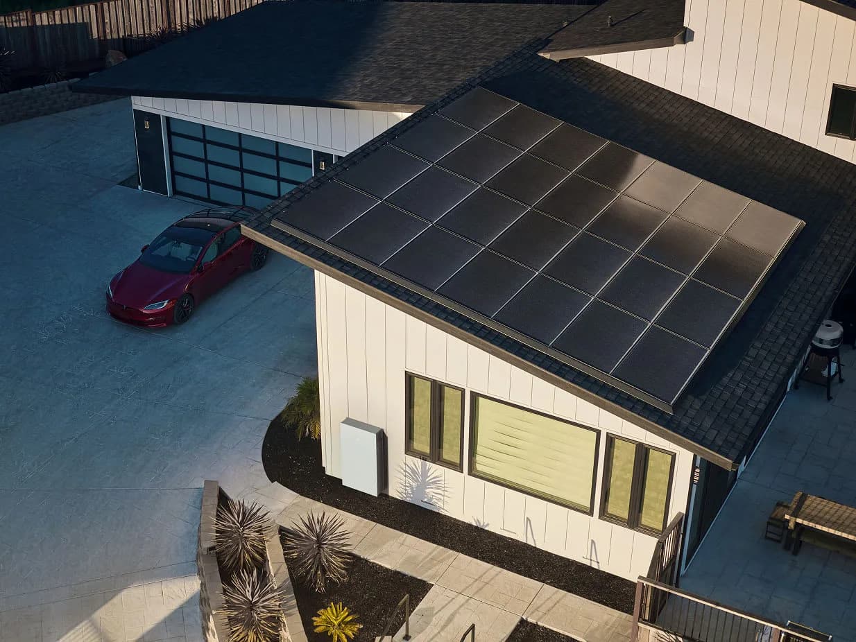 Aerial view of a home with Tesla solar panels on the roof and a wall-mounted Powerwall by the entrance